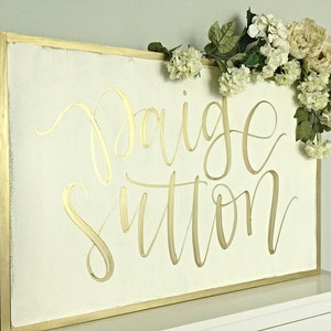 May include: A white wooden sign with a gold frame and the name "Paige Sutton" written in gold calligraphy. The sign is decorated with a garland of white flowers.