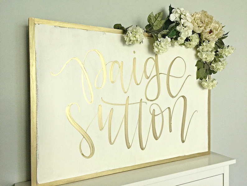 May include: A white wooden sign with a gold frame and the name "Paige Sutton" written in gold calligraphy. The sign is decorated with a garland of white flowers.