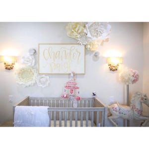 May include: A white cot with a white blanket and a pink and white mobile hanging above it. The wall behind the cot is decorated with white paper flowers and a sign that says "Chandler Paige" in gold lettering.