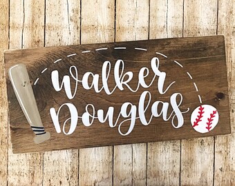 Puede incluir: Un letrero de madera con los nombres "Walker Douglas" escritos en cursiva blanca. Un bate de béisbol y una pelota de béisbol están pintados en el letrero.