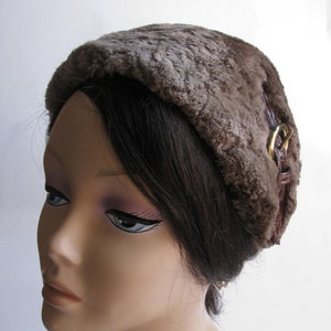 Può includere: Un cappello di pelliccia sintetica marrone con un cinturino in pelle e una fibbia dorata. Il cappello è indossato da un manichino con i capelli castani.