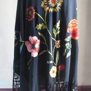 Puede incluir: Chal negro con diseño floral bordado. El chal presenta un gran girasol, flores rojas y rosas y hojas verdes. El borde inferior tiene una larga franja negra.