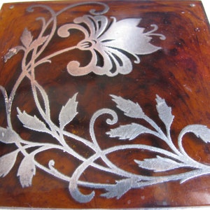 May include: A square wooden box with a silver floral design. The design features a large flower with leaves and vines.