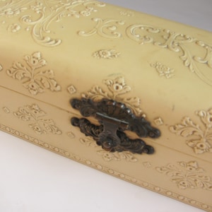 Op de afbeelding: Een crèmekleurige decoratieve doos met een sierlijk reliëf en een metalen sluiting.