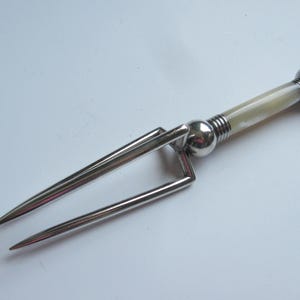 Könnte beinhalten: Eine Vintage-Tranchiergabel in Silberoptik mit cremefarbenem Griff. Die Gabel hat zwei Zinken und einen dekorativen Griff mit abgerundetem Ende. Der Griff hat ein poliertes, perlmuttartiges Aussehen, und die Metallkomponenten sind glänzend.