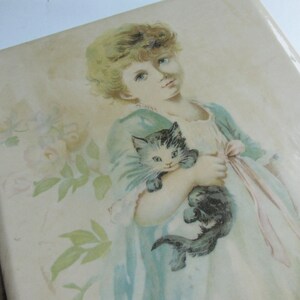 May include: A vintage photo album with a delicate floral design and a young girl holding a black and white kitten. The album has a brass clasp.
