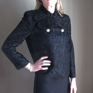 May include: A black textured jacket featuring a double-breasted design with two silver buttons. The jacket has long sleeves and a distinctive pattern. This garment is part of a formal wear collection.
