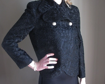 May include: Black textured jacket with a double-breasted design and two silver-colored buttons. The jacket has long sleeves and a unique pattern. The garment is part of a formal wear collection.