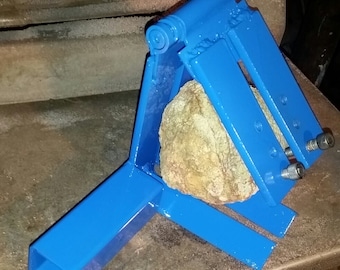 Puede incluir: Una herramienta de división de rocas de metal azul con una roca sujeta en las mordazas. La herramienta tiene una bisagra y un brazo de palanca.