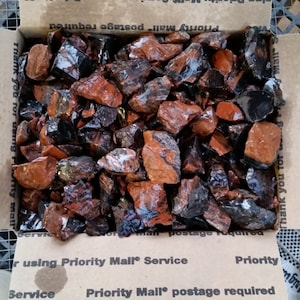 May include: A box filled with red and black striped jasper stones. The stones are rough and have a natural, unpolished look.