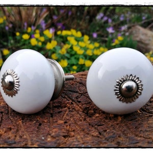 Könnte beinhalten: Zwei weiße Keramik-Schrankknöpfe mit silbernen Akzenten. Die Knöpfe sind rund und haben ein erhabenes, dekoratives Design in der Mitte.