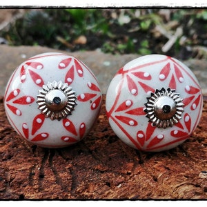 Könnte beinhalten: Zwei weiße Keramik-Schrankknöpfe mit rotem und weißem Blumenmuster. Die Knöpfe haben eine silberne Metallbasis.