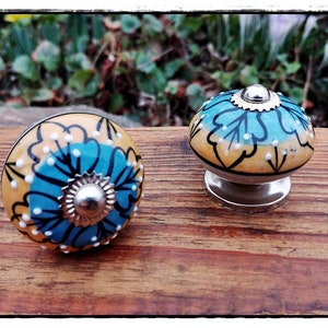 Könnte beinhalten: Zwei Keramik-Schubladengriffe mit einem blauen, gelben und weißen Blumendesign. Die Griffe haben eine silberne Metallbasis.