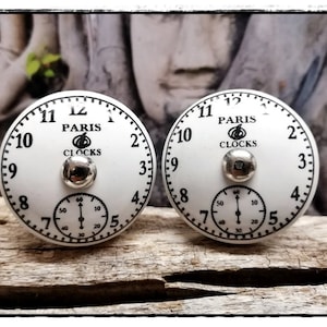 Könnte beinhalten: Zwei weiße Keramik-Uhrenknöpfe mit schwarzen Zahlen und Zeigern. Die Knöpfe haben eine silberne Mitte mit einem kleineren Zifferblatt und dem Text "Paris Clocks" mit einem Uhrensymbol.