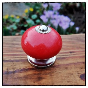 Könnte beinhalten: Roter Keramik-Schrankknopf mit einem silbernen Metallsockel und einem dekorativen silbernen Metallring um die Oberseite.