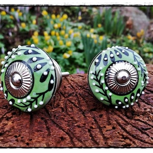 Könnte beinhalten: Zwei grüne Keramik-Schrankknöpfe mit einem schwarzen und weißen Blumendesign und einer silbernen Mitte. Die Knöpfe befinden sich auf einer braunen Holzoberfläche.