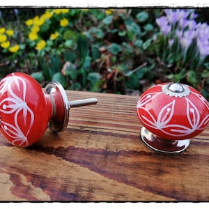 Könnte beinhalten: Zwei rote Keramik-Schrankknöpfe mit weißen Blumenmustern. Die Knöpfe haben eine silberne Metallbasis und eine Schraube.
