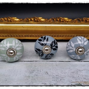 Könnte beinhalten: Drei dekorative Keramik-Schrankknöpfe. Die Knöpfe sind rund mit einer silbernen Metallbasis. Einer ist hellgrün mit weißem Blumendesign, einer ist hellblau mit schwarzem Blumendesign und einer ist hellblau mit weißem Blumendesign.