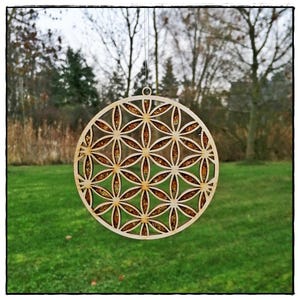 Könnte beinhalten: Runder Holzschmuck mit einem lasergeschnittenen Lebensblumenmuster. Der Schmuck wird an einem dünnen Faden aufgehängt und hat eine goldfarbene Glitzerfüllung in den ausgeschnittenen Formen. Der Hintergrund zeigt grünes Gras und Bäume, leicht unscharf.