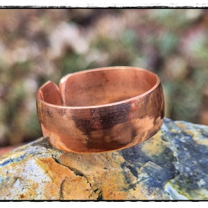 Könnte beinhalten: Ein Kupferring mit geteiltem Design, der auf einem strukturierten Felsen ruht. Der Ring hat ein breites Band und eine warme, rötlich-braune Farbe. Der Felsen hat eine Mischung aus Blau-, Gelb- und Brauntönen und bietet einen natürlichen Hintergrund.