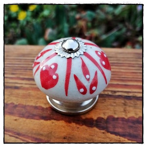 Op de afbeelding: Witte keramische kastknop met een rood en wit bloemenmotief. De knop heeft een zilveren metalen basis en een zilveren metalen bovenkant.