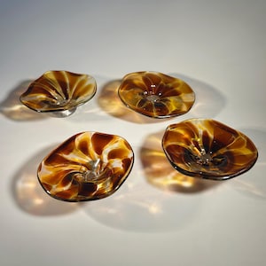 May include: Four decorative glass bowls with a unique, organic shape. The bowls feature a swirl design in shades of amber and brown, with a clear center. Each bowl is elevated on a small, silver-colored base, creating a floating effect.