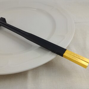 May include: A pair of black chopsticks with gold tips. The chopsticks are resting on a white plate.