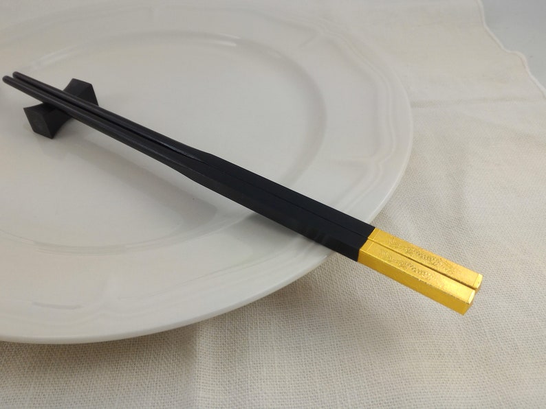 May include: A pair of black chopsticks with gold tips. The chopsticks are resting on a white plate.