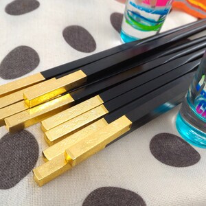 May include: A set of black chopsticks with gold tips. The chopsticks are laid out on a white surface with black polka dots. The chopsticks have a decorative pattern on the gold tips.