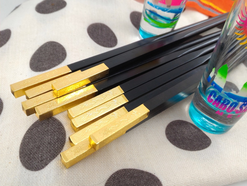 May include: A set of black chopsticks with gold tips. The chopsticks are laid out on a white surface with black polka dots. The chopsticks have a decorative pattern on the gold tips.