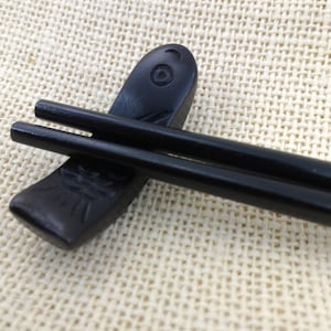 May include: Black chopsticks with a fish-shaped wooden rest. The rest has a carved design of a fish.