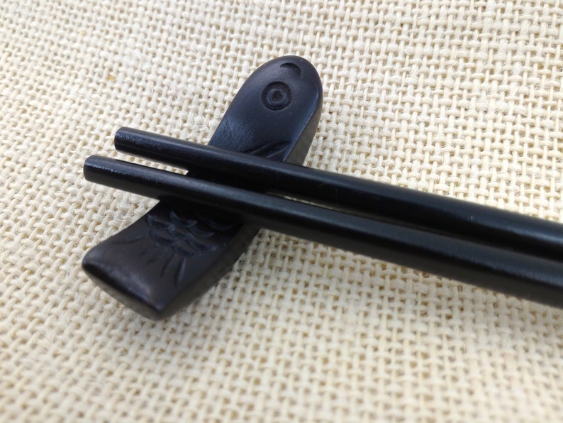 May include: Black chopsticks with a fish-shaped wooden rest. The rest has a carved design of a fish.