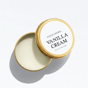 Op de afbeelding: Open, goudkleurige blikken doos met vaste Vanilla Cream parfum. Het deksel is eraf en onthult het romige, gebroken witte product. Het deksel heeft een wit etiket met de woorden "COCO + REBEL" en "VANILLA CREAM SOLID PERFUME" in zwarte tekst.