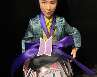 May include: A doll wearing a teal and pink patterned jacket, yellow shirt, and purple ribbon. The doll is holding a clear ornament filled with small cards. A purple logo is attached to the ornament.