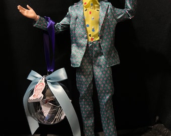 May include: A doll wearing a teal suit with a pink and teal geometric pattern, a yellow shirt with colorful buttons, and white high-top sneakers. The doll is holding a clear ornament with a blue ribbon.
