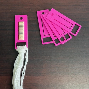 May include: A set of bright pink plastic embroidery floss bobbins. One bobbin is wrapped with white embroidery thread and features a paper label with the number 3865 and a barcode. The other four bobbins are empty and stacked in a fan shape.
