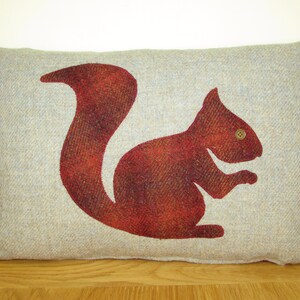 Könnte beinhalten: Ein Kissen aus grauem und weißem Tweed mit einem roten Eichhörnchen-Design. Das Eichhörnchen ist braun umrandet und hat einen Knopf für ein Auge.