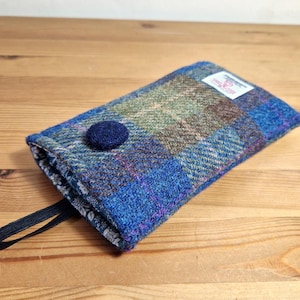 May include: A rectangular, blue, green, and brown plaid fabric case with a dark blue button closure and a black elastic loop. The case has a small white tag and is set on a wooden surface.
