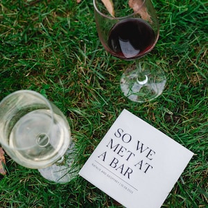 May include: A white card with black text that reads "So we met at a bar" and "Natalie and Geoffrey | 10.08.2023". Two glasses of wine are on the grass next to the card. One glass is filled with red wine and the other with white wine.