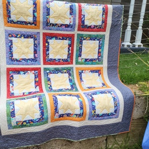 May include: A handmade quilt featuring a repeating pattern of yellow star blocks. Each star is set within a colorful square, with a patterned border in shades of blue, orange, red, and green. The quilt has a gray border and orange binding.