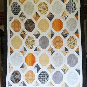 May include: A white quilt with a geometric pattern of octagons and triangles. The octagons are in shades of gray, brown, and yellow, and the triangles are white. The quilt is edged with a black border.