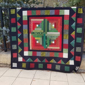 Könnte beinhalten: Eine schwarze, rote und grüne Quiltdecke mit einem Blockhausmuster in der Mitte. Die Decke ist von einem schwarzen Rand mit einem geometrischen Muster umgeben.
