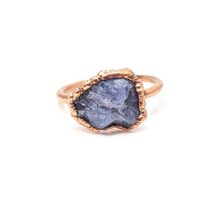 May include: A copper ring with a raw, blue-grey gemstone set in the center. The ring has a textured, hammered finish.