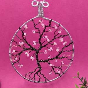 Op de afbeelding: Een draadgewikkelde levensboom-sculptuur met roze kralen die bloemen voorstellen. De sculptuur is rond en hangt aan een zilveren draadlus.
