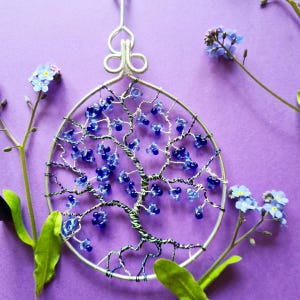 Op de afbeelding: Een draadgewikkelde levensboom met blauwe glazen kralen, hangend tegen een paarse achtergrond. De boom is omringd door vergeet-me-nietjes.