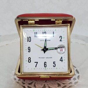 May include: A vintage Phinney-Walker travel alarm clock. The clock has a white face with black numerals and hands, and a red case. The clock face reads "DATE-ALARM" and "PHINNEY-WALKER". The clock is open and ready to use.