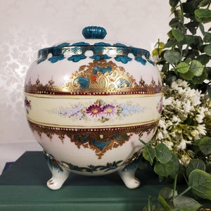 May include: A white, lidded ceramic jar with a teal and gold floral design. The jar has a round shape with three small feet. The lid is topped with a teal knob. The jar is decorated with a band of flowers and gold accents.