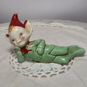 May include: A ceramic elf figurine in a reclining pose, featuring a glossy green outfit and a red pointed hat. The elf has a pale face with rosy cheeks, blue eyes, and pointed ears. There is visible paint wear on the red hat and collar.