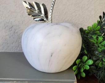 May include: A decorative white marble apple with gray veining and a silver leaf and stem. The apple is sitting on a gray book with the text "d Summers DOUBLEDAY". The apple is a decorative item.