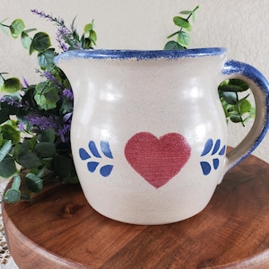May include: Cream-colored ceramic pitcher with a blue rim and handle. A red heart is painted on the front, flanked by blue floral accents. The pitcher is likely used for serving beverages and has a rustic, handmade aesthetic.